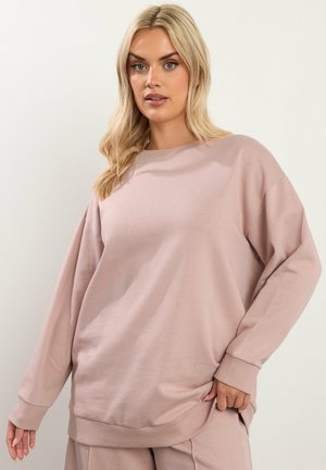 Vrouw met lang blond haar, gekleed in een losse, licht mauve sweatshirt en bijpassende broek, staand tegen een eenvoudige lichte achtergrond.