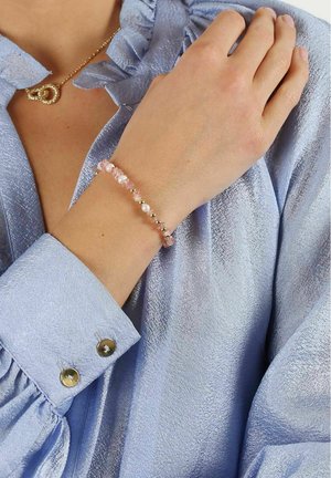 Beaded bracelet made of pink crystals, white pearls, and gold accents. Worn on a wrist, paired with a light blue textured blouse.