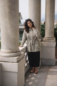 A woman wears a beige animal-print jacket with a cinched waist and a black skirt. She stands against stone pillars, with greenery in the background.