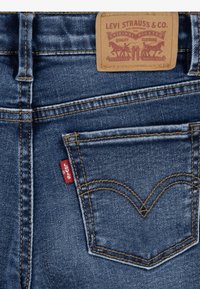 Plave traperice s stražnjim džepom i kožnom etiketom Levi Strauss & Co. te crvenom Levi's tekstilnom oznakom na rubu džepa.