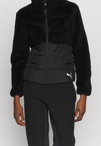 Zwarte jas met een vachtachtig bovenstuk en een gladde, gevoerde onderkant. Ritssluiting, hoge kraag en klein wit logo aan de voorkant.