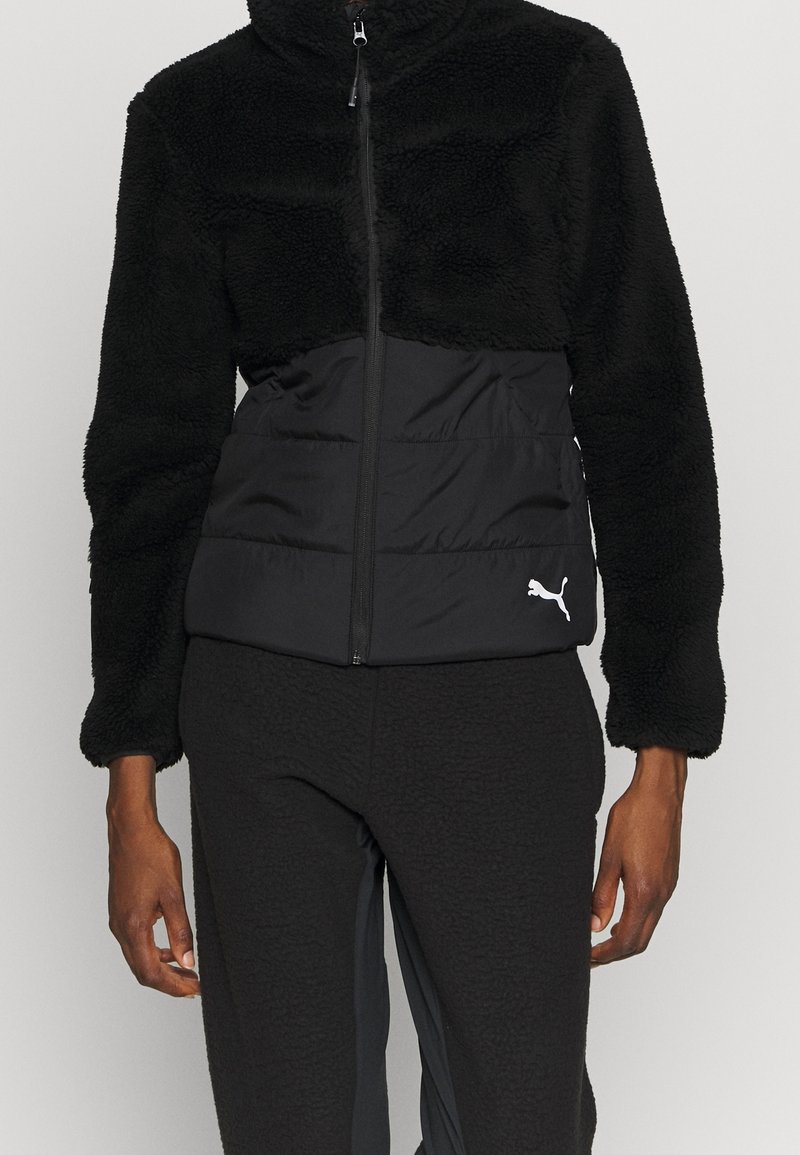 Zwarte jas met een vachtachtig bovenstuk en een gladde, gevoerde onderkant. Ritssluiting, hoge kraag en klein wit logo aan de voorkant.