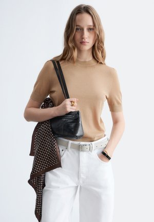 Žena s vlnitými vlasmi má na sebe béžové tričko s krátkym rukávom a biele nohavice, drží čiernu kabelku so vzorovanou šatkou cez rameno, ruka v vrecku.