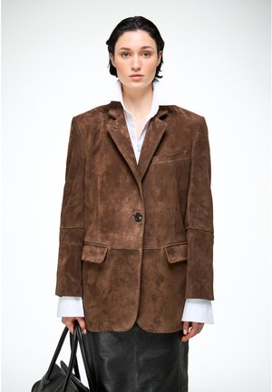 Jonge vrouw met kort donker haar, gekleed in een oversized bruine suède blazer, wit overhemd, zwarte leren rok, en ze draagt een zwarte handtas.