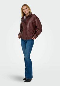 Giacca in pelle borgogna con vestibilità rilassata, caratterizzata da un colletto alto, due tasche frontali e orlo a coste. Abbinata a jeans a zampa d'elefante blu.