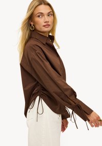 Chemise marron à boutons avec fronces sur les côtés et détails noués aux poignets, confectionnée dans un tissu léger, associée à un pantalon blanc.