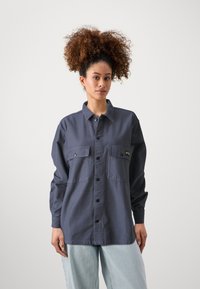 Dark blue button-up shirt with two chest pockets, long sleeves, and a relaxed fit, worn over light blue jeans. Soft, textured fabric.
