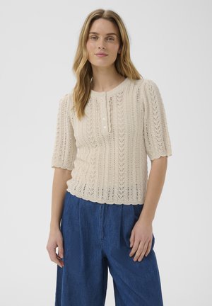 Frau mit langen blonden Haaren, die ein beiges kurzärmeliges Strickoberteil mit Knöpfen und hoch taillierte, lockere blaue Hose trägt, steht vor einem einfarbigen Hintergrund.