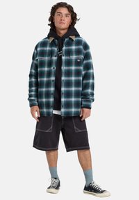 Geruite flanellen overhemd in teal en zwart, met een knoopsluiting, gecombineerd met een zwarte hoodie en donkere shorts. Zwarte sneakers en blauwe sokken.