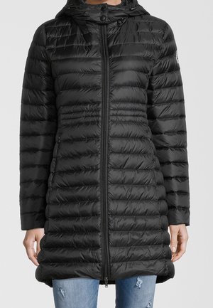 Schwarze, gesteppte Steppjacke mit Kapuze und Frontreißverschluss, getragen über zerrissenen blauen Jeans, Hände seitlich locker hängend.