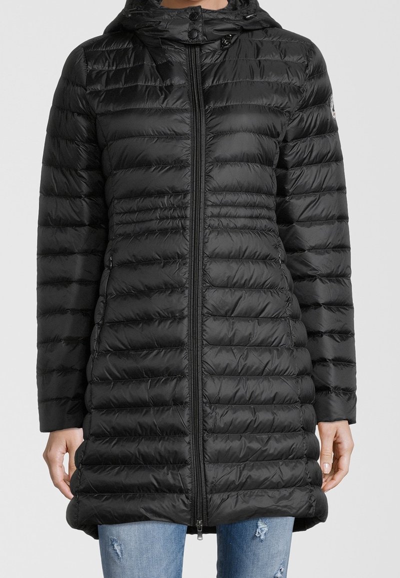 Sort quiltet dunjakke med hætte og frontlynlås, båret over revnede blå jeans, hænder hvilende ved siden.