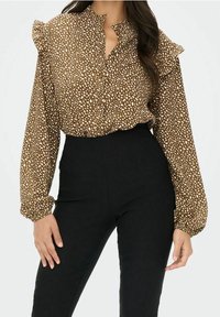 Blouse marron avec un motif tacheté blanc, col froncé et manches longues, associée à un pantalon noir taille haute. Texture de tissu lisse.