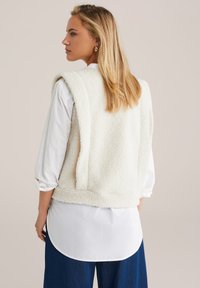 Kremno obarvan, teksturiran flis brezrokavnik z zasnovo spuščene rame, nosen čez belo srajco s gumbi, kar prikazuje večplastni videz.