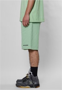 Pantaloni corti di cotone verde chiaro con una vestibilità ampia e il logo "S N' I M SENSE." sull'orlo, abbinati a sneakers grigie e calze nere alla caviglia.