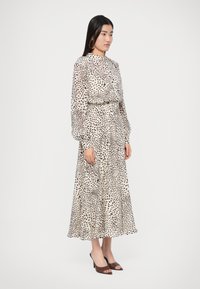 Hosszú, áttetsző bézs ruha fekete pettyes mintával, hosszú ujjakkal, magas nyakkivágással és deréknál összeszűkítve. Barna, nyitott orrú magassarkúval párosítva.