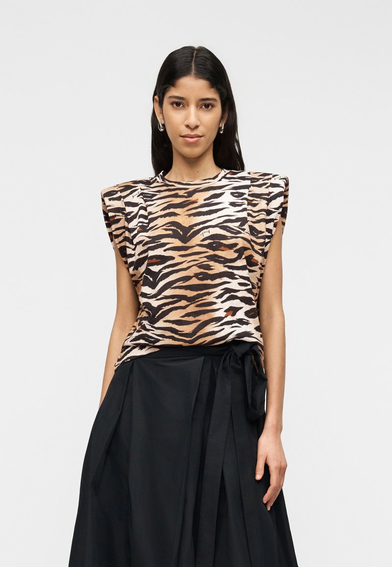 Γυναίκα με μακριά σκουρόχρωμα μαλλιά, που φοράει αμάνικη μπλούζα με σχέδιο τίγρης και επενδεδυμένους ώμους, συνδυασμένη με ψηλόμεση μαύρη φούστα δεμένη στη μέση.