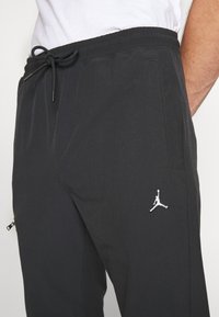 Schwarze Sporthose mit elastischem Bund, Kordelzugverschluss, Seitentaschen und einem weißen Jumpman-Logo-Akzent am linken Oberschenkel.