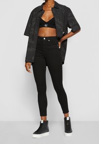 Persona con una camiseta corta negra de Calvin Klein, una camisa de botones negra oversize con logo, jeans skinny negros y zapatillas altas negras.