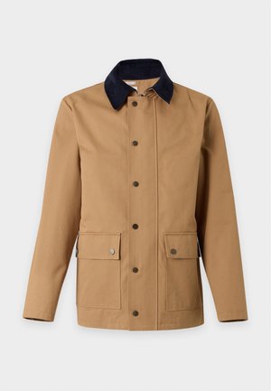 Jeune homme portant une veste beige, une chemise noire boutonnée, un pantalon gris ample et des baskets beiges, debout avec une main dans la poche sur fond blanc.