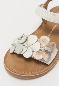 Sandalia de cuero blanco con plantilla de gamuza marrón, que presenta tres apliques de flores en capas en blanco, plateado y dorado metálico, acentuados con perlas.