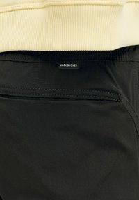 Gros plan d’un pantalon noir avec une poche arrière et une petite étiquette noire indiquant « JACK & JONES » sous un pull côtelé crème.