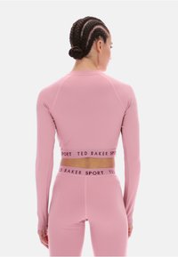 Rózsaszín, hosszú ujjú sportfelső, amely cropped fazonú, texturált derékrésszel rendelkezik, amelyen a "TED BAKER SPORT" márkajelzés kontrasztos színben található.