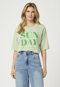 Šviesiai žalia per didelė marškinė su tamsiai žalia "SUN DAY" grafika, trumpomis rankovėmis ir apvaliu iškirpimu, derinama su aukštu juosmeniu mėlynomis džinsomis.