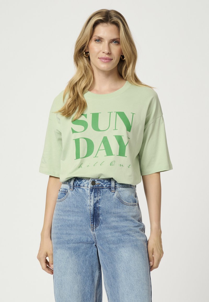 Šviesiai žalia per didelė marškinė su tamsiai žalia "SUN DAY" grafika, trumpomis rankovėmis ir apvaliu iškirpimu, derinama su aukštu juosmeniu mėlynomis džinsomis.