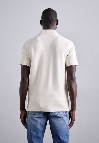 Polo krekls off-white krāsā, tekstilā adīta materiālā, īsām piedurknēm un ar klasisku apkakli, apvienots ar gaiši ziliem džinsiem.