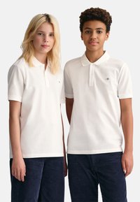 Chemises polo blanches à manches courtes et patte de boutonnage, portées avec un pantalon en velours côtelé foncé. Emblème discret sur chaque chemise.