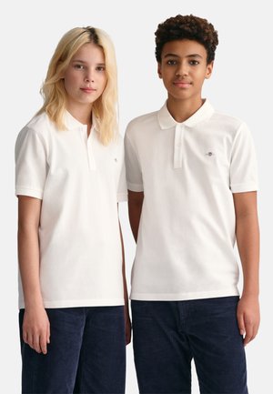 Chemises polo blanches à manches courtes et patte de boutonnage, portées avec un pantalon en velours côtelé foncé. Emblème discret sur chaque chemise.