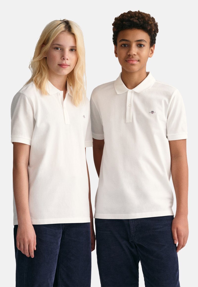 Chemises polo blanches à manches courtes et patte de boutonnage, portées avec un pantalon en velours côtelé foncé. Emblème discret sur chaque chemise.