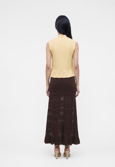 Femme aux longs cheveux noirs portant un haut jaune sans manches et une jupe longue en dentelle marron, debout de dos sur fond blanc.