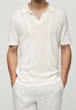 Mann trägt ein kurzärmeliges, cremefarbenes Strick-Poloshirt und helle Hose mit den Händen in den Taschen.