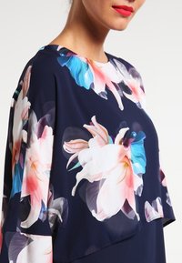 Květinový top v námořnické tkanině, zdobený velkými růžovými a modrými květy. Volný střih s kulatým výstřihem a krátkými rukávy. Měkká textura.