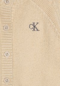 Cardigan bej tricotat, cu un guler cu nervuri, cinci nasturi și un mic logo brodat "cK" în albastru închis.