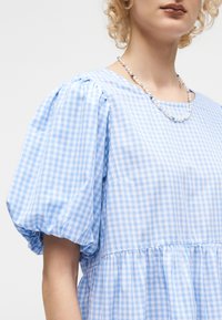 Personne aux cheveux bouclés portant une robe à carreaux bleue et blanche à manches bouffantes et un collier de perles et de perles bleues.