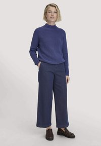 Frau steht mit den Händen in den Taschen, trägt einen blauen Rippensweater, weit geschnittene dunkelblaue Hosen und dunkelbraune Loafers vor einem hellen Hintergrund.