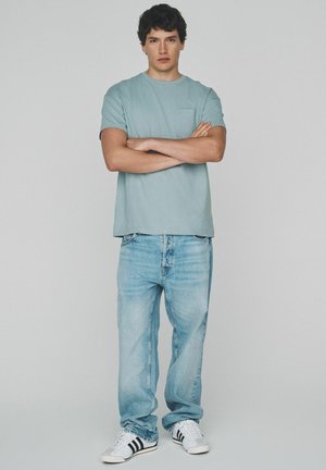 Noor mees seisab käed risti, seljas helesinine T-särk, lahtised tuhmunud teksad ja valged tossud mustade triipudega, ühtlase taustaga.