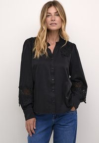 Camisa negra de botones con un escote rizado, detalles de encaje en los hombros y las mangas, y una textura suave parecida a la satén. Corte casual.
