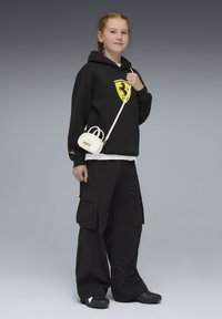 Sweat à capuche noir avec un logo Ferrari, associé à un pantalon cargo noir ample. Porte un petit sac à main blanc avec un logo Puma. Chaussures noires texturées.