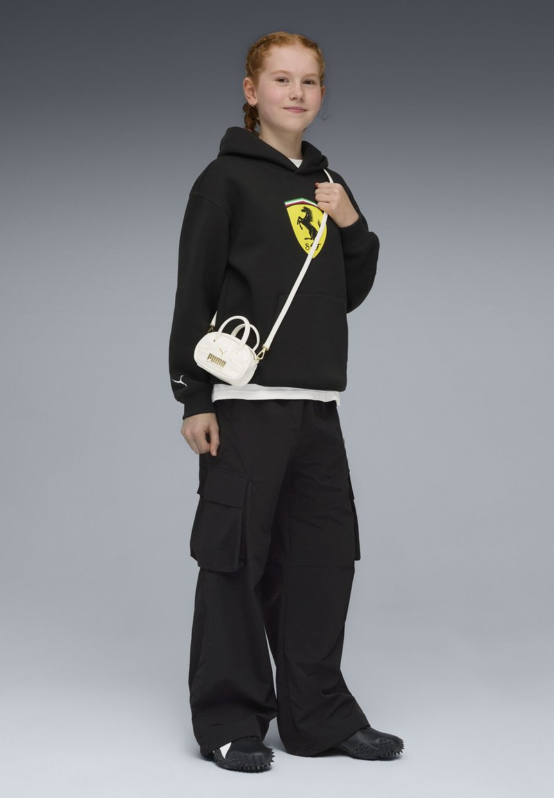 Sweat à capuche noir avec un logo Ferrari, associé à un pantalon cargo noir ample. Porte un petit sac à main blanc avec un logo Puma. Chaussures noires texturées.