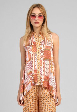 Femme portant un chemisier sans manches à motif patchwork et un pantalon assorti à motifs, avec des lunettes de soleil teintées rose, debout devant un fond gris uni.