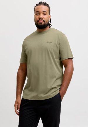 Mand med flettet hår og skæg iført en olivengrøn t-shirt og sorte bukser, stående med den ene hånd i lommen mod en ensfarvet baggrund.