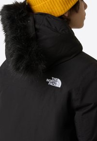 Μαύρο μονωμένο μπουφάν με τελείωμα κουκούλας από συνθετική γούνα, με λευκό λογότυπο στο στήθος και υφασμένο κίτρινο σκουφί.