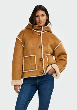 Modèle portant une veste en peau lainée couleur camel, avec des bordures crème, de gros boutons et des poches plaquées, associée à un jean bleu sur un fond uni.