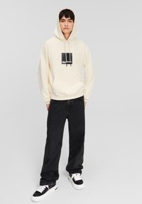 Felpa con cappuccio crema con un logo grafico nero sul petto, abbinata a jeans neri larghi e sneakers bianco e nere.