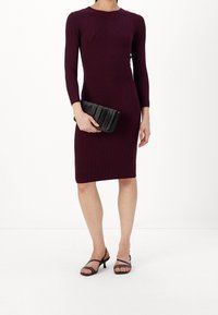 Abito a coste bordeaux con maniche lunghe e forma aderente. Una pochette nera arricciata e tacchi neri con cinture completano l'outfit.