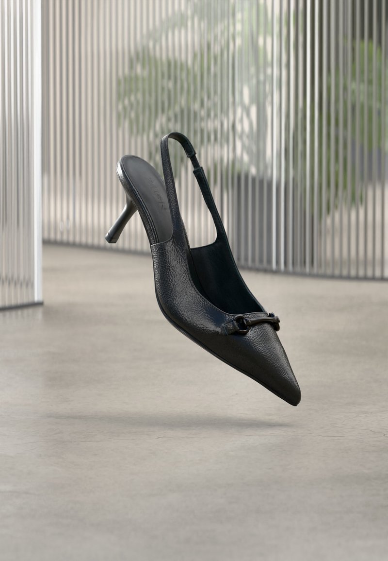 Chaussure à talon haut à bride arrière en cuir noir texturé avec un bout pointu et une petite boucle décorative, flottant au-dessus d'un sol lisse.
