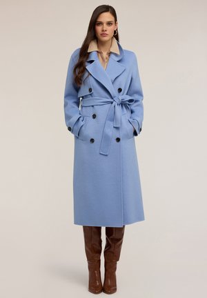 Cappotto lungo blu in misto lana, a doppio petto con bottoni neri, vita con cintura, ampie rever e tasche laterali. Abbinato a stivali marroni al ginocchio.